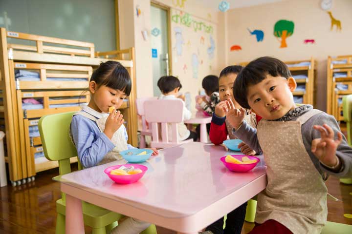 Children sitting at a daycare table eating snacks in a bright, cheerful room with animal-themed wall art.